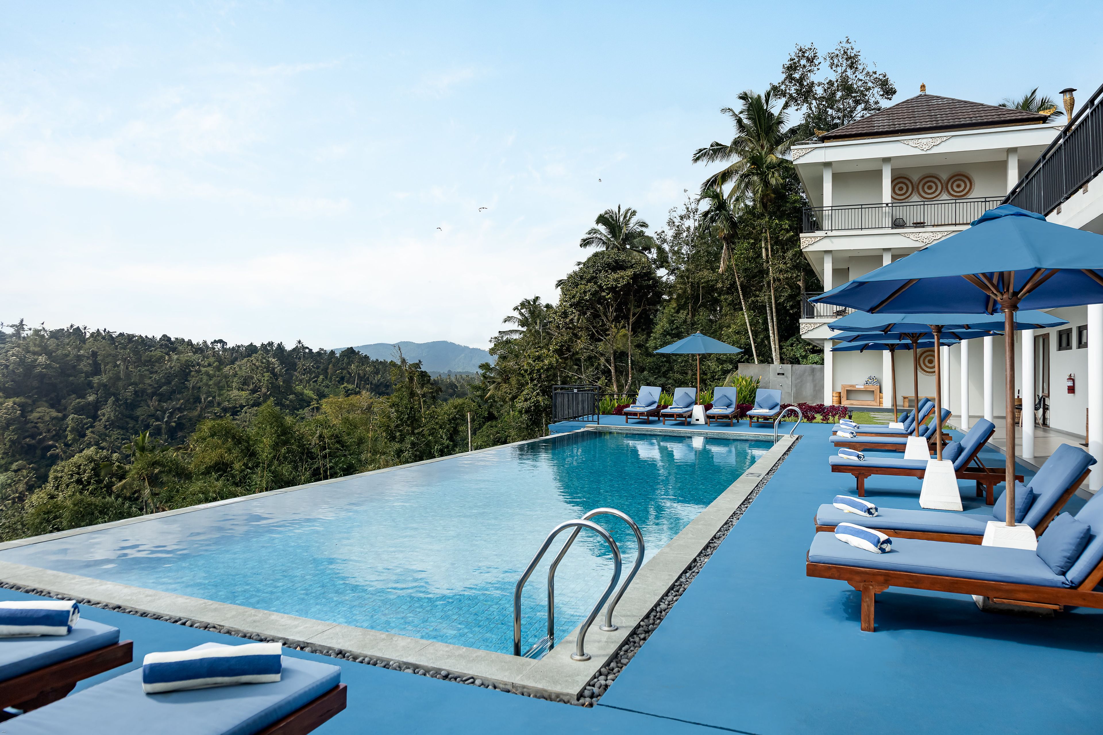 A poolside area with lounge chairs and umbrellas, surrounded by lush greenery and a view of distant mountains.
