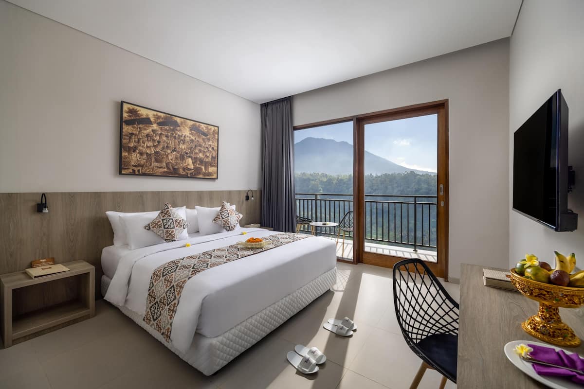 A hotel room with a large bed, a desk with a fruit bowl, a TV, and a balcony with a mountain view.