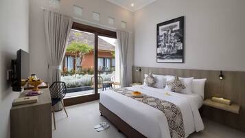 A modern hotel room with a large bed, a desk with a computer, a chair, and a view of a garden through the window.