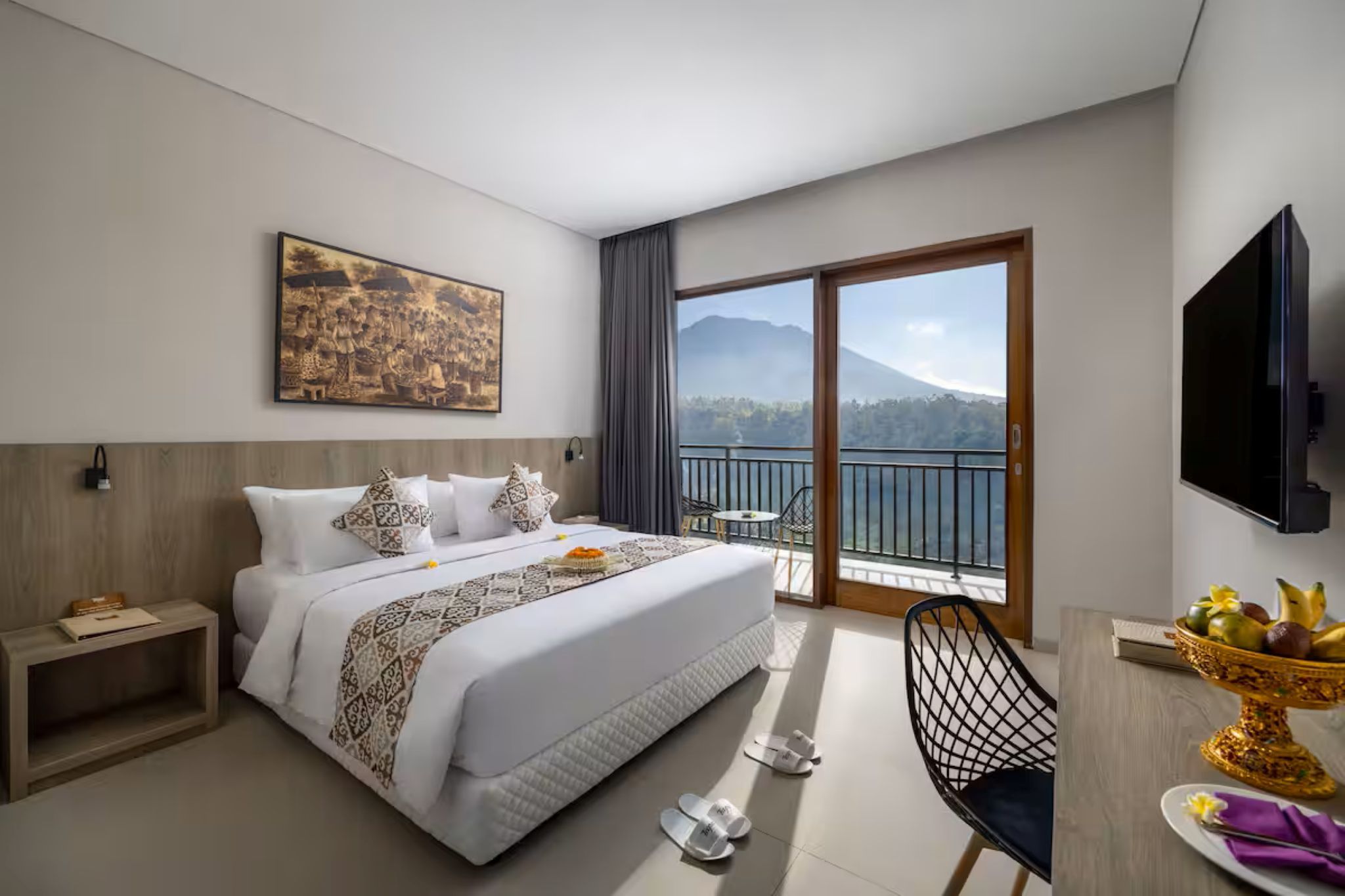 A hotel room with a bed, a dining table with fruit, a TV, and a balcony with a mountain view.