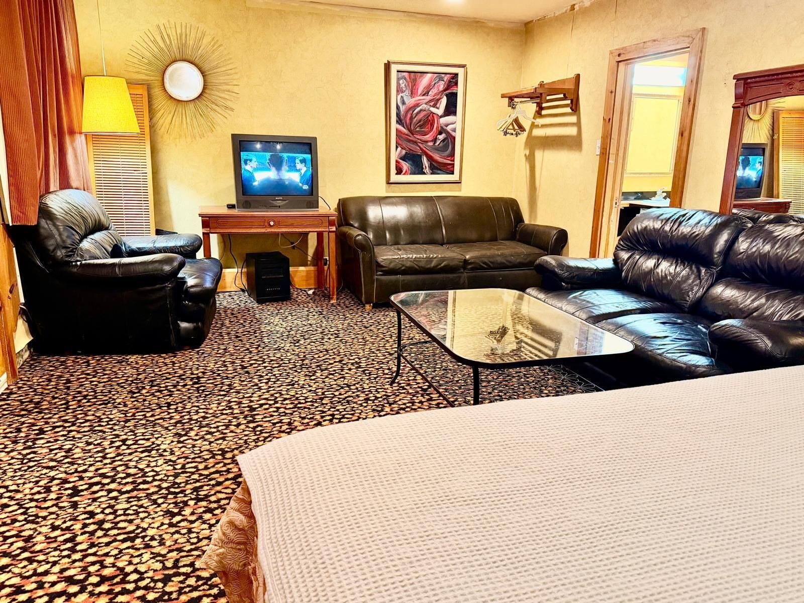A hotel room with a black leather sofa, a glass coffee table, a television on a wooden stand, and a patterned carpet.