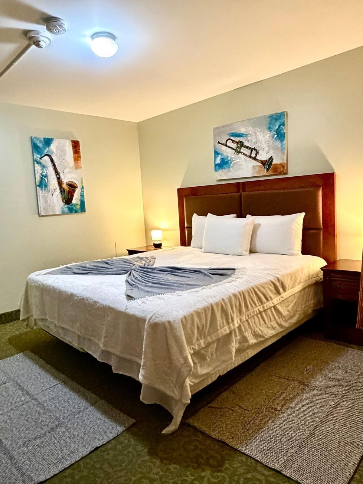 Habitación de hotel con una cama grande, dos cuadros en las paredes, un ventilador de techo y una mesita de noche con lámpara.