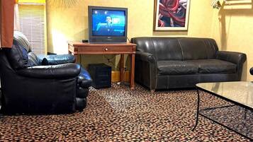 A living room with a patterned carpet, a black leather sofa, a glass coffee table, a television on a wooden stand, and a wall-mounted decorative plate.