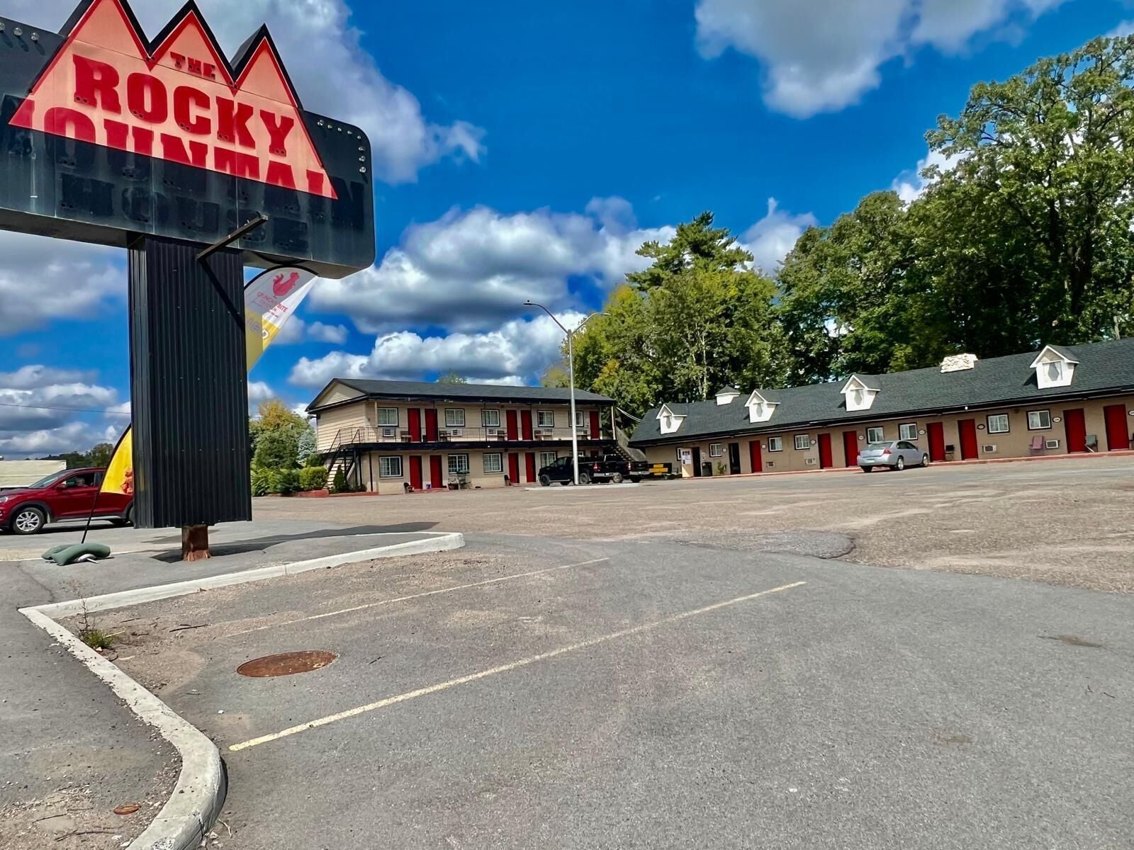 A motel with a sign reading "The Rocky Mountain." It features a two-story building with multiple rooms, a parking lot, and a clear blue sky with clouds.