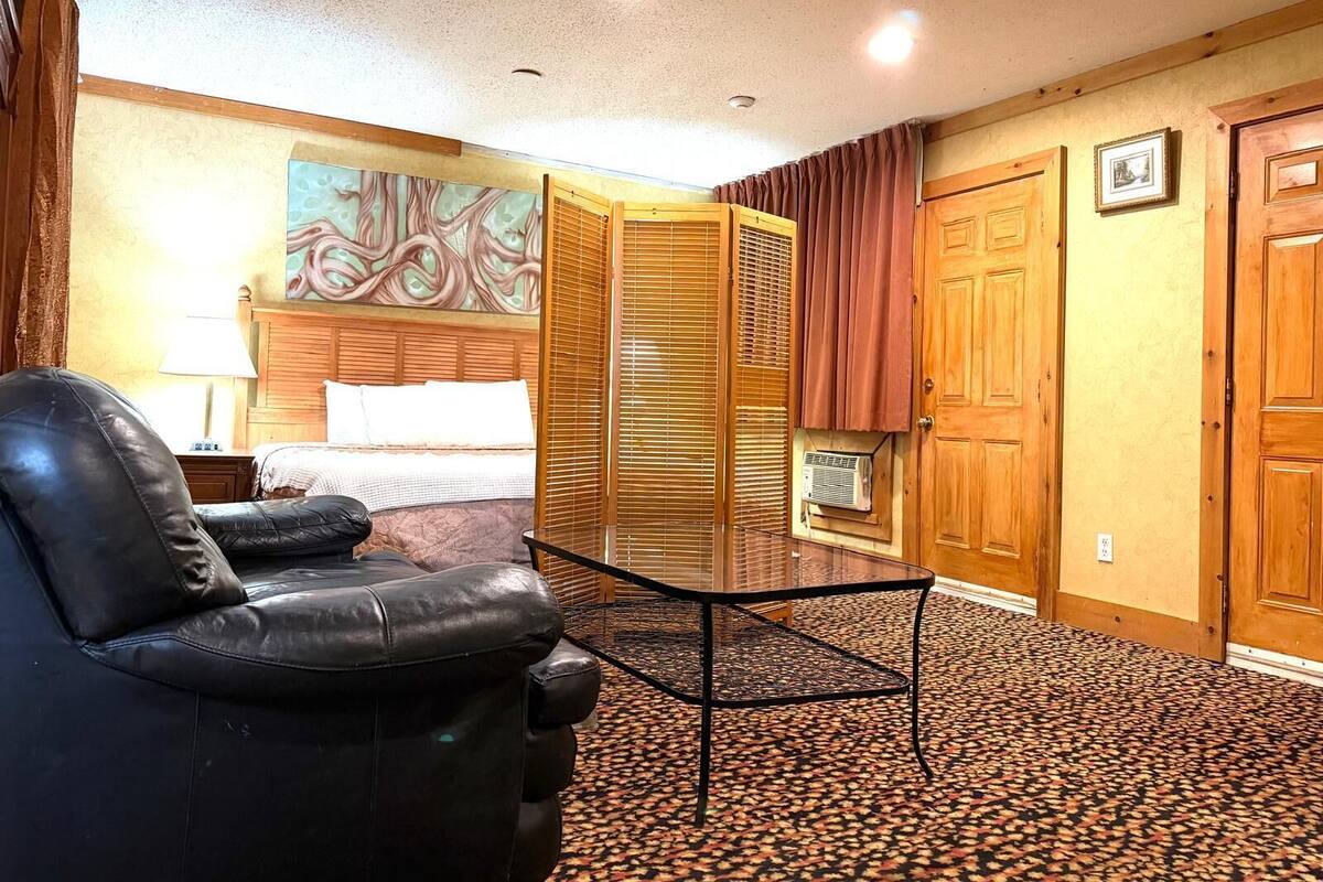 A hotel room with a bed, a black leather chair, a glass table, and a patterned carpet.