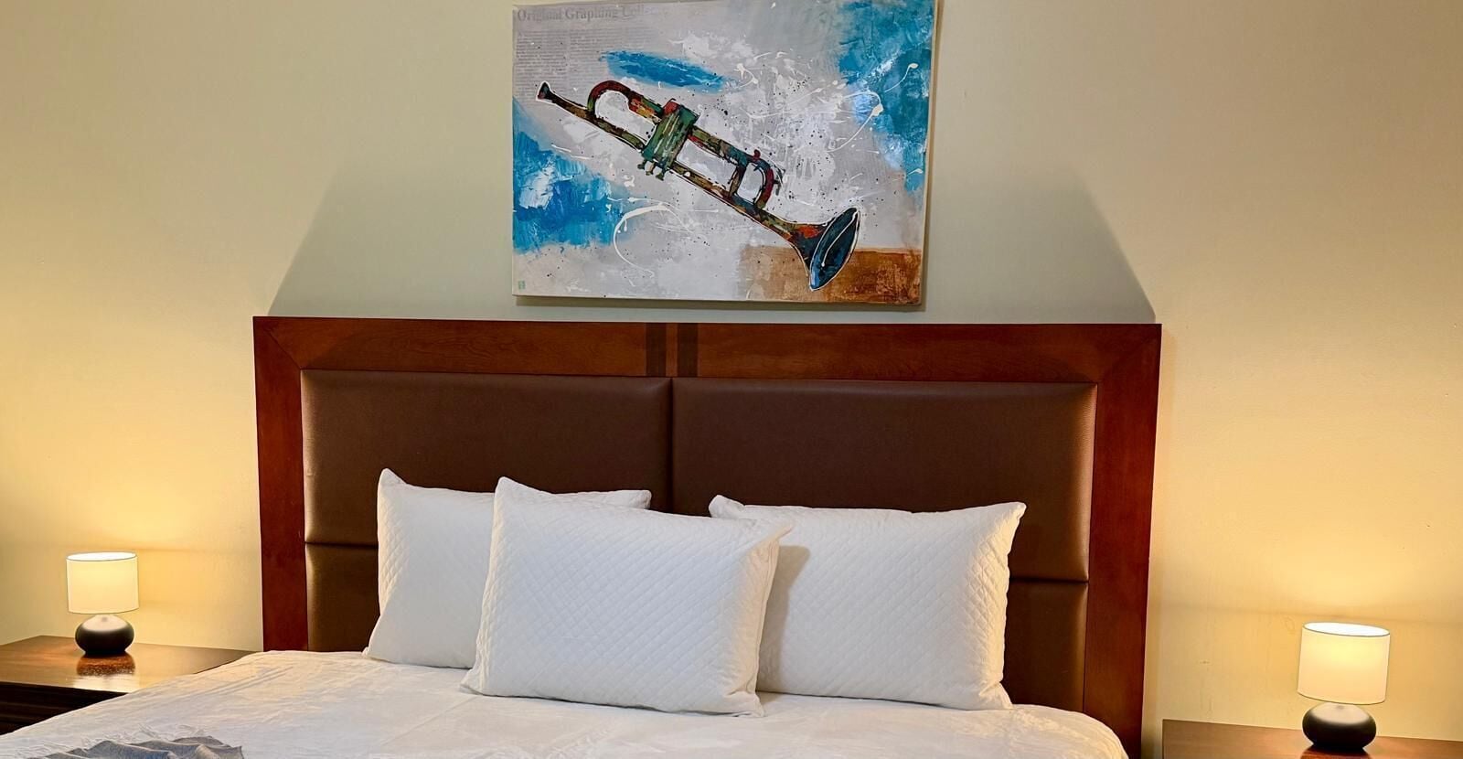 A hotel room with a large bed, two white pillows, a wooden headboard, and a painting of a trumpet on the wall.