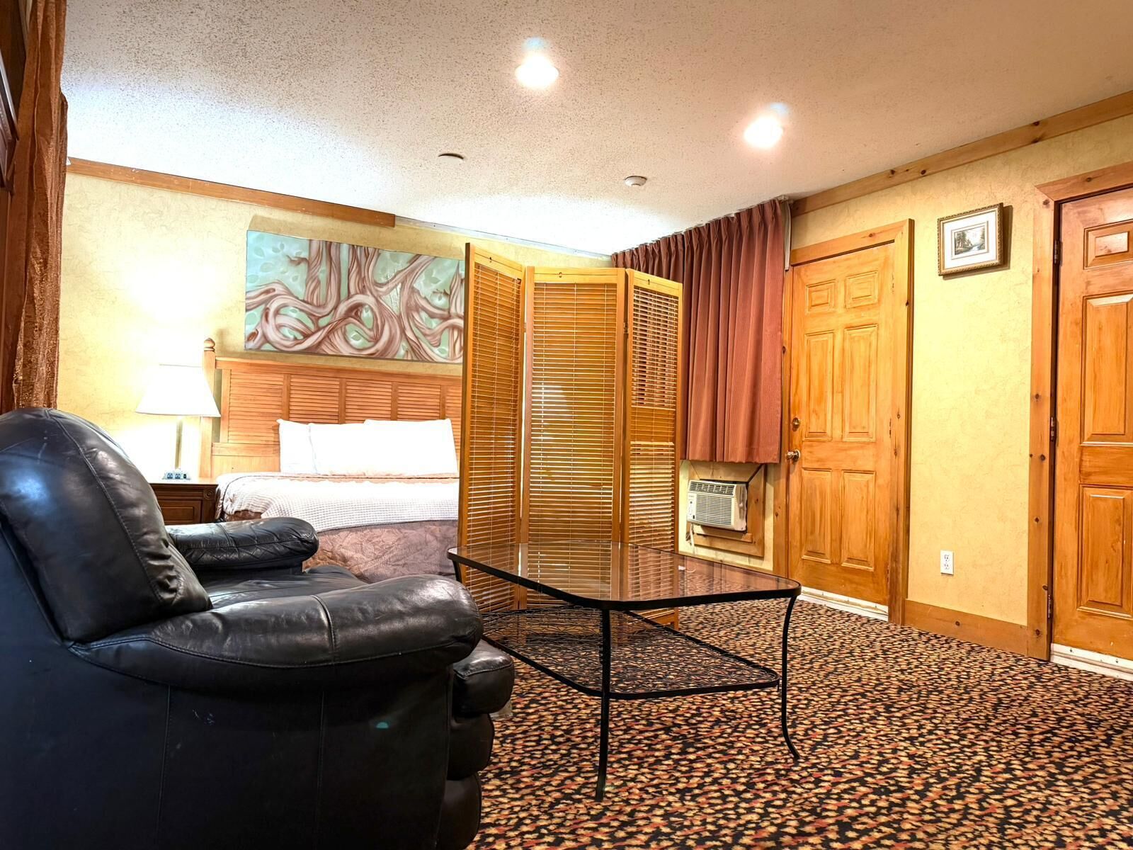 Habitación de hotel con cama, un sillón de cuero negro, una mesita de vidrio, un armario de madera y un cuadro en la pared.