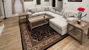 Un salon moderne avec un canapé d’angle, une table basse en bois, un tapis à motifs, un lustre et un tableau accroché au mur.