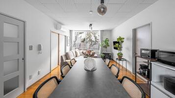 A modern dining area with a long table, chairs, a vase of flowers, and a kitchen area with appliances.