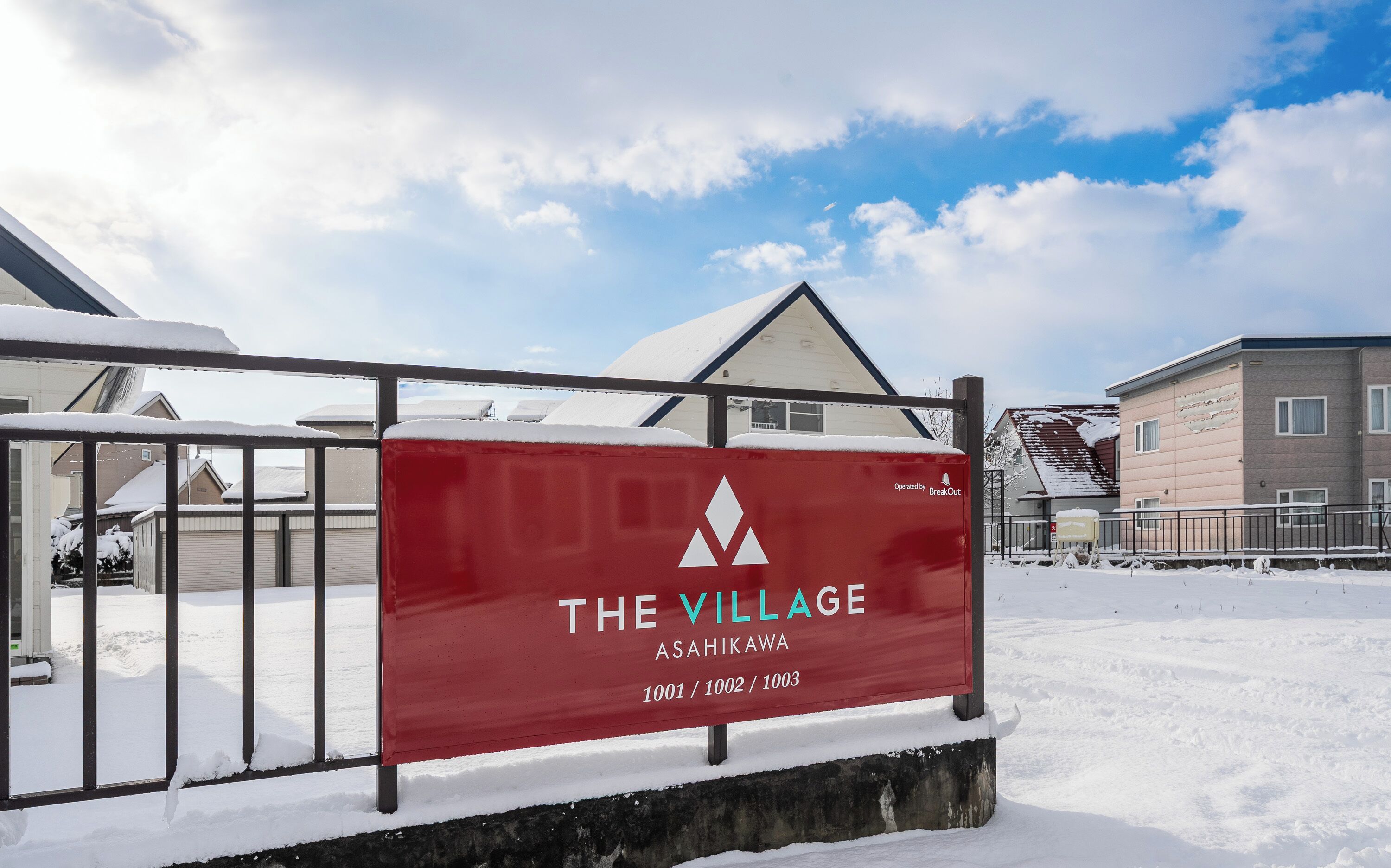 A sign for "The Village Asahikawa" with a red background and white lettering, indicating the location of a residential area.