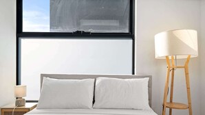 A bedroom with a large window, a bed with white bedding, a floor lamp, and a small bedside table with a lamp. - One-Bed Apartment Close to Cafés and Coast (Port Melbourne)