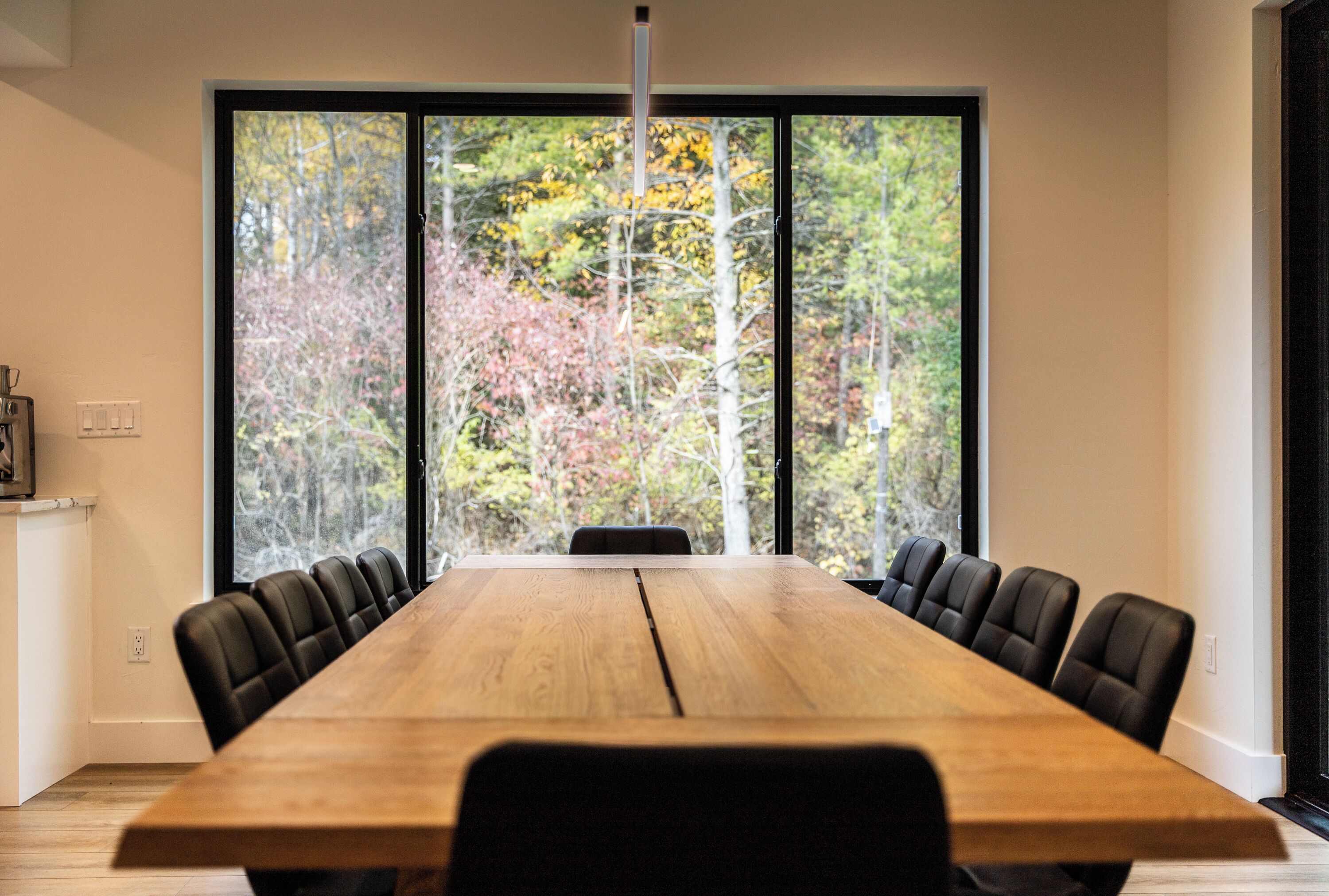 Une salle de conférence avec une longue table en bois et des chaises noires, de grandes fenêtres donnant sur la forêt.