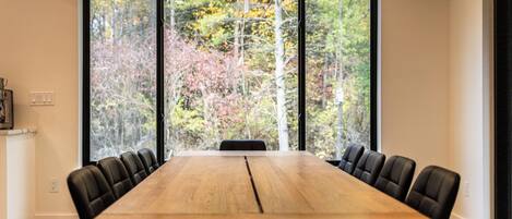 Une salle de conférence avec une longue table en bois et des chaises noires, de grandes fenêtres donnant sur la forêt.