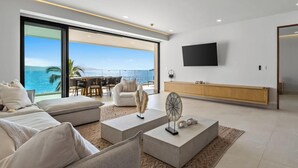 A modern living room with a large window overlooking the sea, a flat-screen TV mounted on the wall, and a minimalist wooden cabinet. - Panarea 3D | Beachfront 4BR + Ocean Views & Pools (Bucerías)