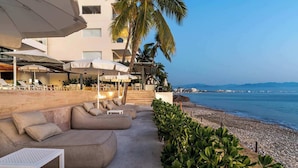 A beachside lounge with outdoor seating and umbrellas, overlooking a pebble beach and the sea. - 4BR w/ Oceanfront Views, Gym & Pools | Panarea 5D (Bucerías)