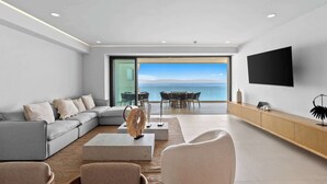 Ein modernes Wohnzimmer mit grauem Sofa, Couchtisch und Fernseher an der Wand. Es gibt einen Essbereich mit Tisch und Stühlen sowie Meerblick durch ein großes Fenster.