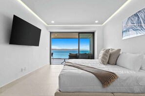 A modern bedroom with a large bed, a flat-screen TV, and a balcony with a sea view.
