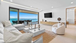 A modern living room with a large window overlooking the sea, a flat-screen TV mounted on the wall, and a minimalist design. - Panarea 4D | Beachfront 4BR + Elevated Ocean Views (Bucerías)