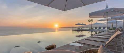 Una piscina infinita con sillones de descanso y sombrillas, con vistas al océano al atardecer.