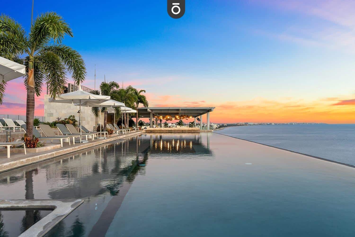 Piscina infinita com vista para o oceano ao pôr do sol, cadeiras de descanso e palmeiras.
