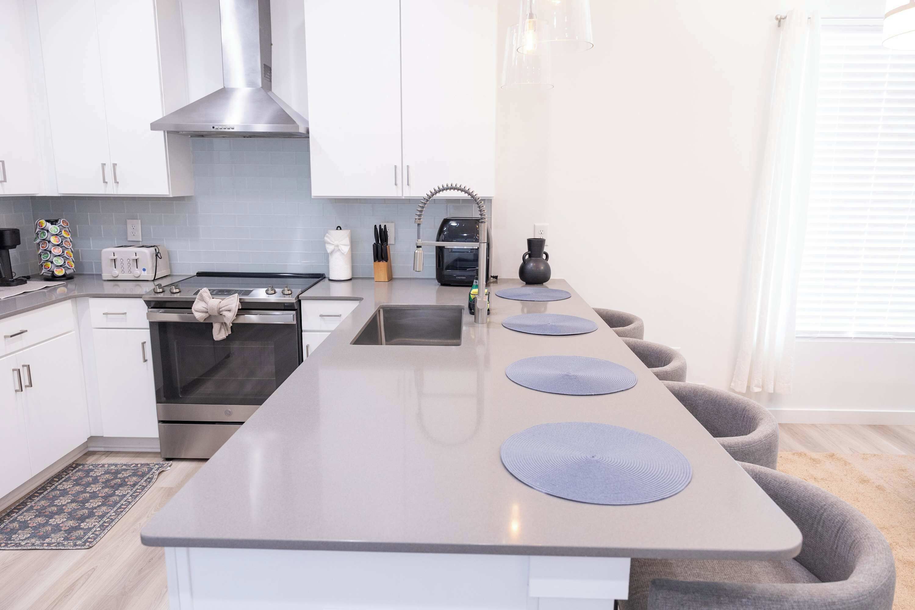 A modern kitchen with white cabinets, a stainless steel range hood, a built-in oven, a toaster, and a sink. There are three grey cushioned seating areas along the counter.