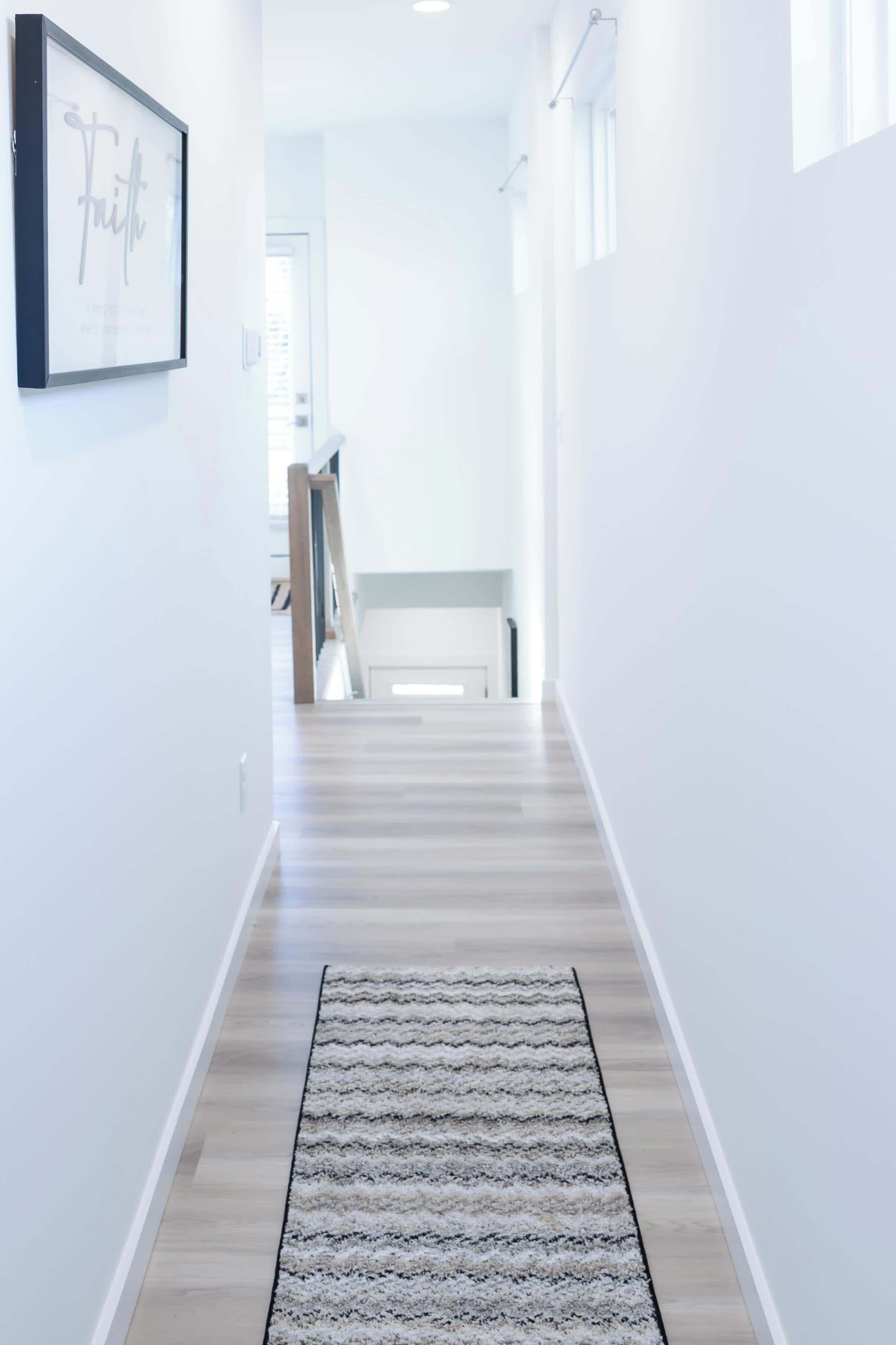 A hallway with a light-colored wooden floor, a patterned rug, and a framed sign on the wall.