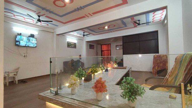 A modern hotel lobby with a glass display table featuring potted plants, a ceiling with geometric patterns, and a television mounted on the wall. - Shubh Hotel (Phulpur)