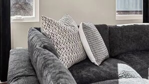 Un salon moderne avec un canapé gris, des coussins à motifs et une vue sur la ville grâce à de grandes fenêtres.