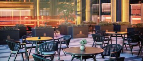 An outdoor seating area with round tables and chairs, potted plants, and a checkered floor pattern.