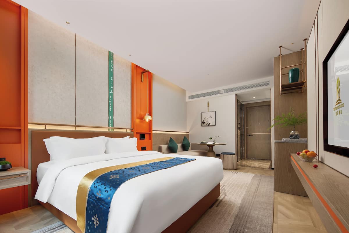 A hotel room with a large bed, a desk, a chair, and a small table with a vase and fruit.