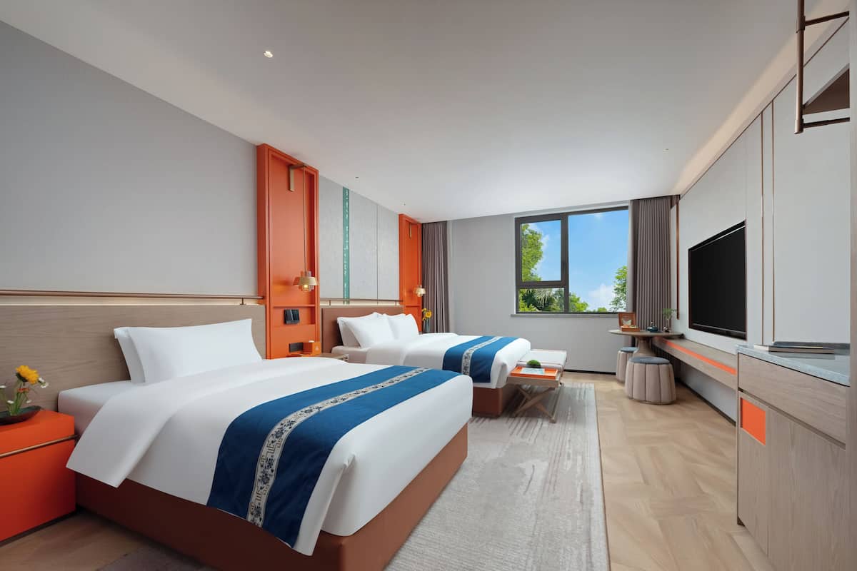 A modern hotel room with two beds, a TV, and a large window offering a view of greenery.