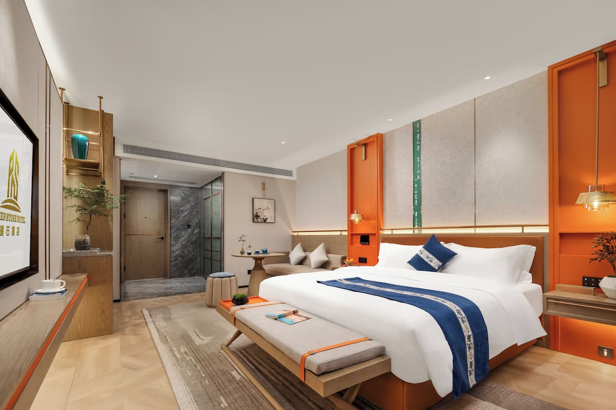 A modern hotel room with a large bed, a bench, a desk, and a reception area.