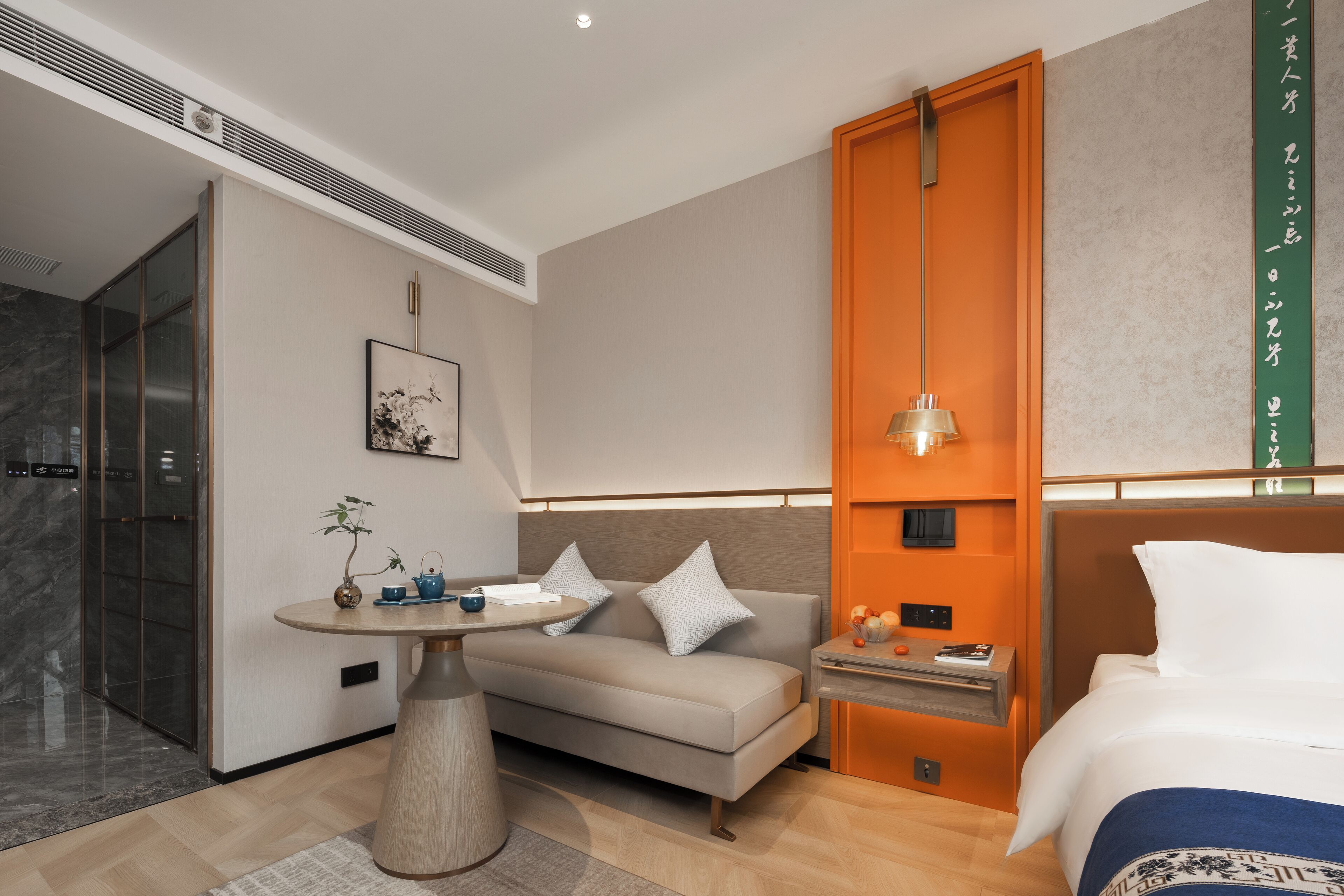 A modern hotel room with a sofa, a small table with a plant, a bedside table with a lamp, and a bed with a blue blanket.