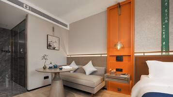 A modern hotel room with a sofa, a small table with a plant, a bedside table with a lamp, and a bed with a blue blanket.