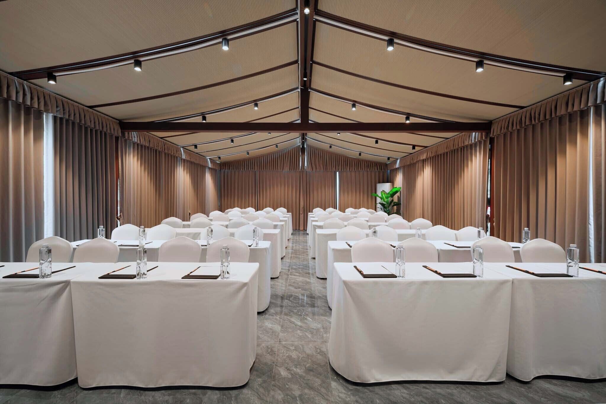 Une salle de conférence avec des rangées de tables, des chaises et un plafond haut doté de luminaires.