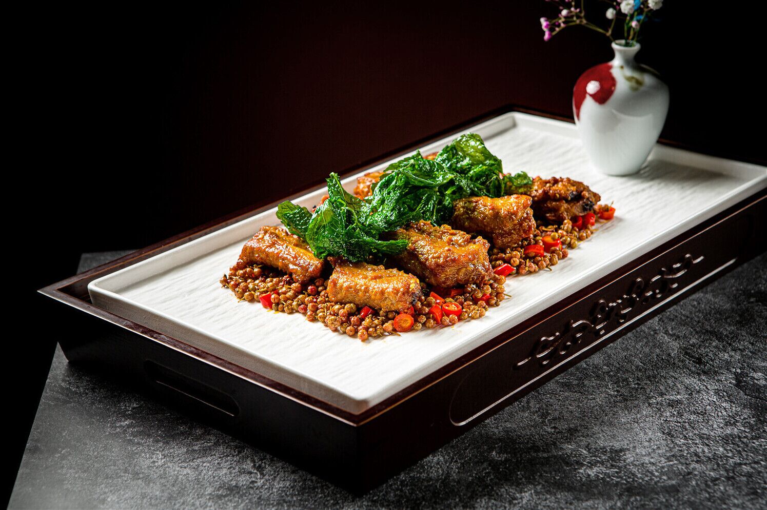 A rectangular plate with a dish of stir-fried meat, vegetables, and peanuts, accompanied by a white vase with flowers.