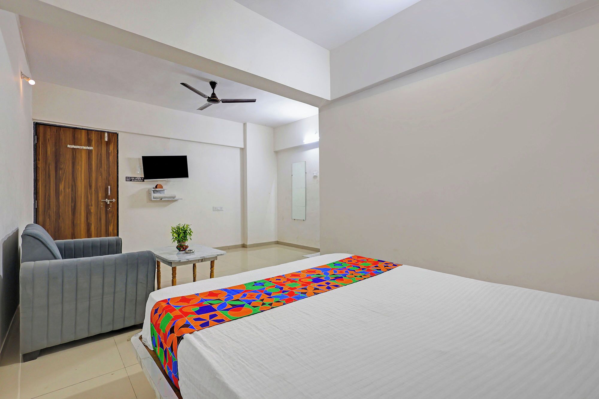 A hotel room with a bed, a sofa, a small table with a plant, a ceiling fan, and a TV mounted on the wall.