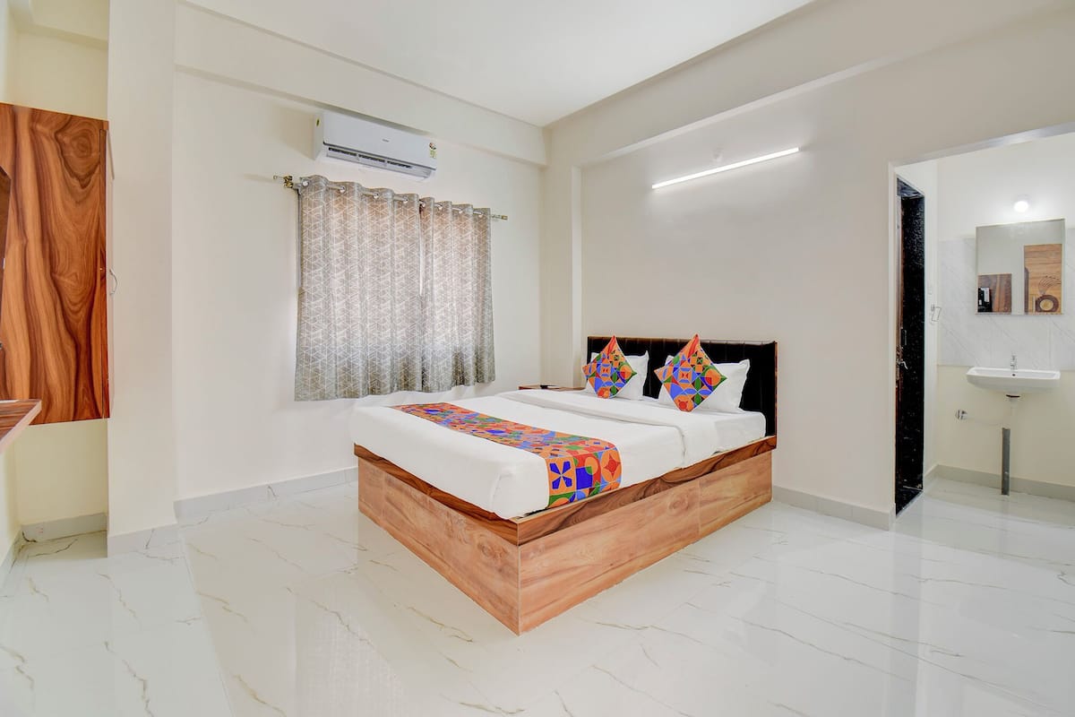 A bedroom with a wooden bed frame, a white mattress, and colorful pillows. There is a window with patterned curtains, an air conditioning unit on the wall, and a bathroom with a sink and mirror.