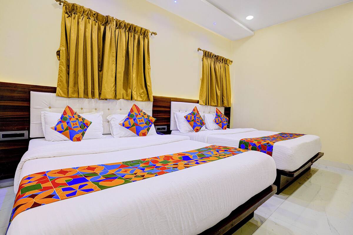 A hotel room with two beds, each with a colorful geometric-patterned bedspread, white bedding, and matching pillows. The room has a wooden headboard, a wooden floor, and yellow curtains.