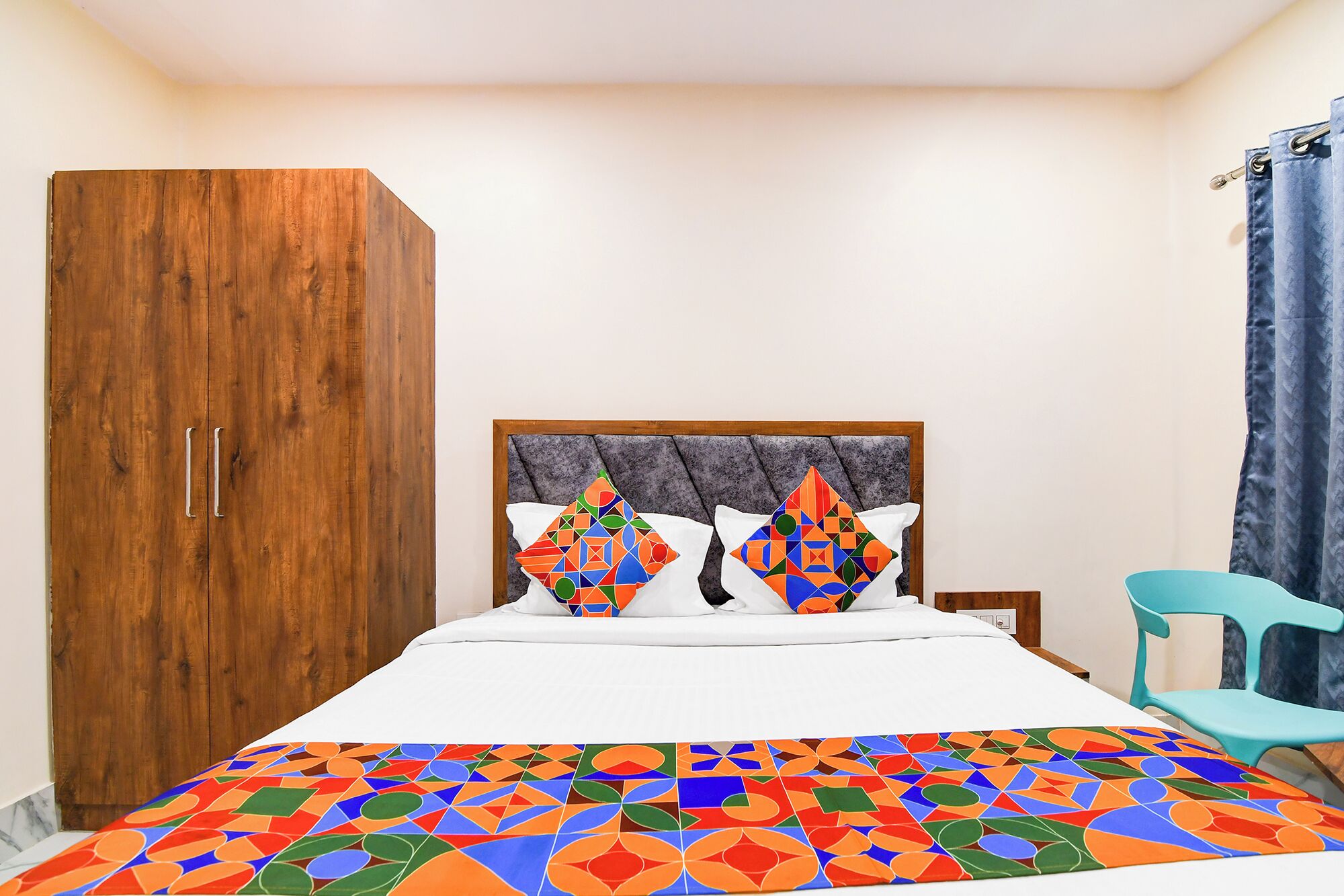A bedroom with a bed, a wooden wardrobe, a blue chair, and a patterned bedspread.