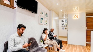 Tres personas sentadas en sillas modernas en una habitación con suelo de madera y paredes blancas, con un televisor montado y fotografías enmarcadas en la pared.