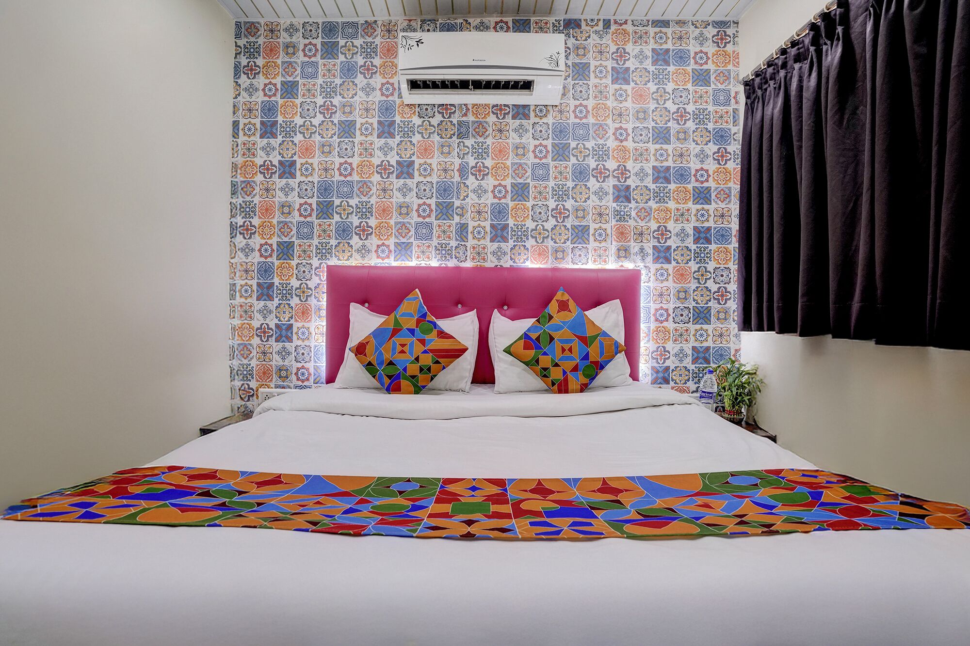 Een slaapkamer met een betegelde muur met patroon, een roze gestoffeerd hoofdeinde, twee kleurrijke kussens en een beddeken met een geometrisch ontwerp.