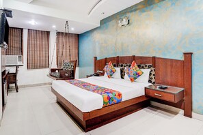 A hotel room with a wooden bed, a nightstand, a desk with an air conditioner, and a window with blinds. - Fabhotel Shivdev International (New Delhi)