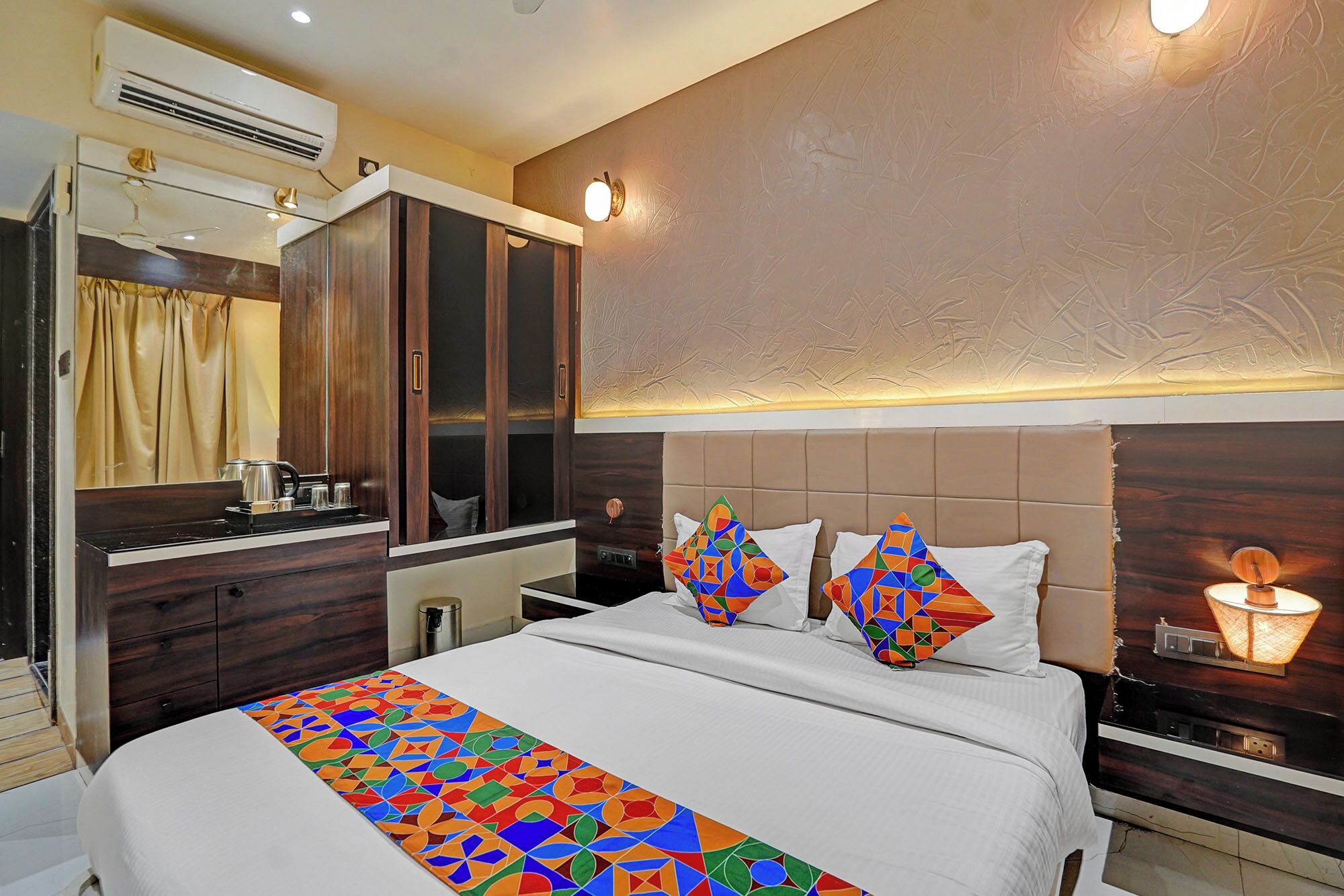 A hotel room with a bed, a wardrobe, a mirror, and a bedside table with a lamp.