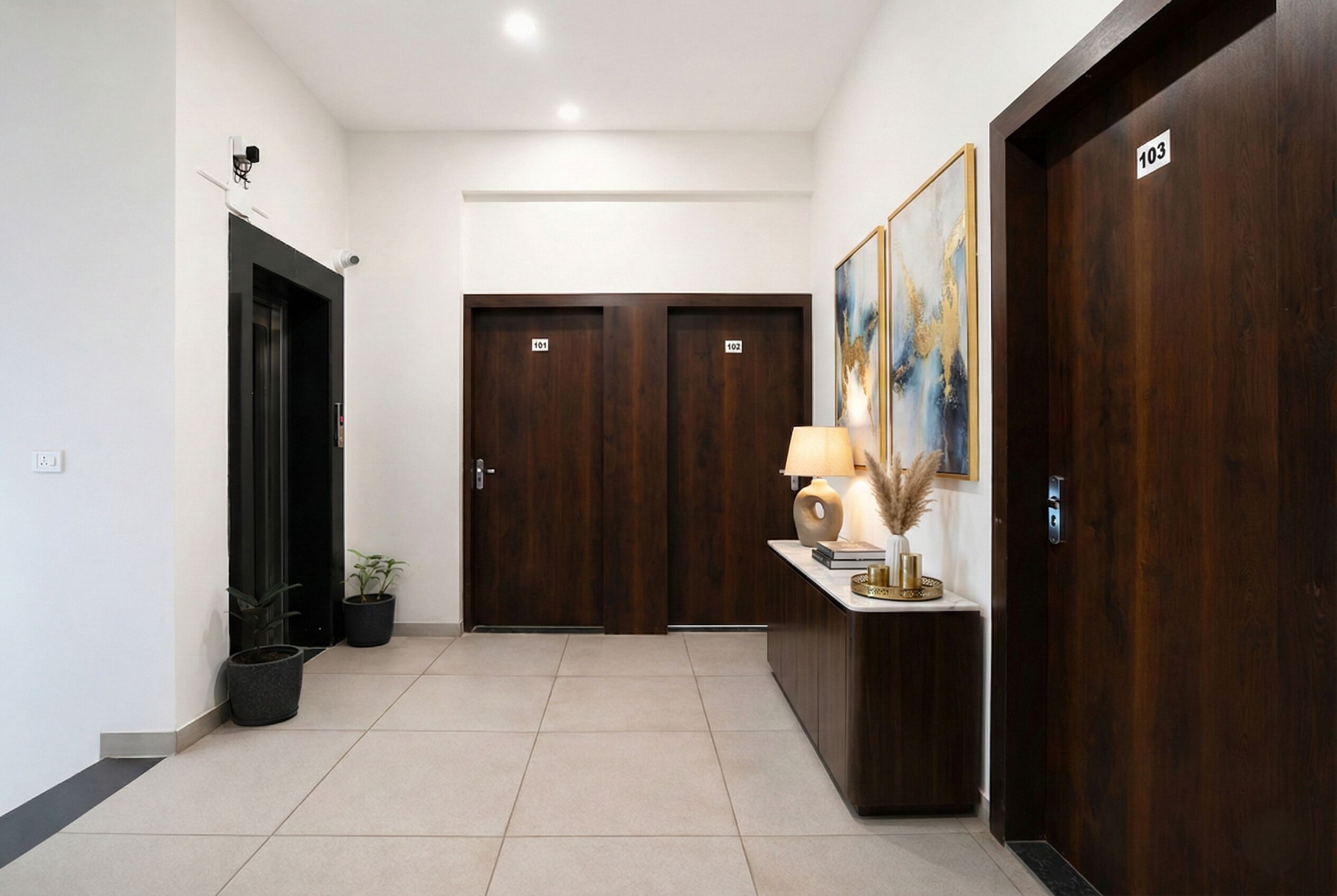 Vestíbulo de hotel con una recepción, dos puertas de madera con las indicaciones 101 y 102, un cuadro en la pared y una planta en maceta.