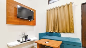 A modern living room with a flat-screen TV mounted on a wooden panel, a blue sofa, a wooden coffee table, and a window with beige curtains. - Fabhotel Olive Inn (Indore)