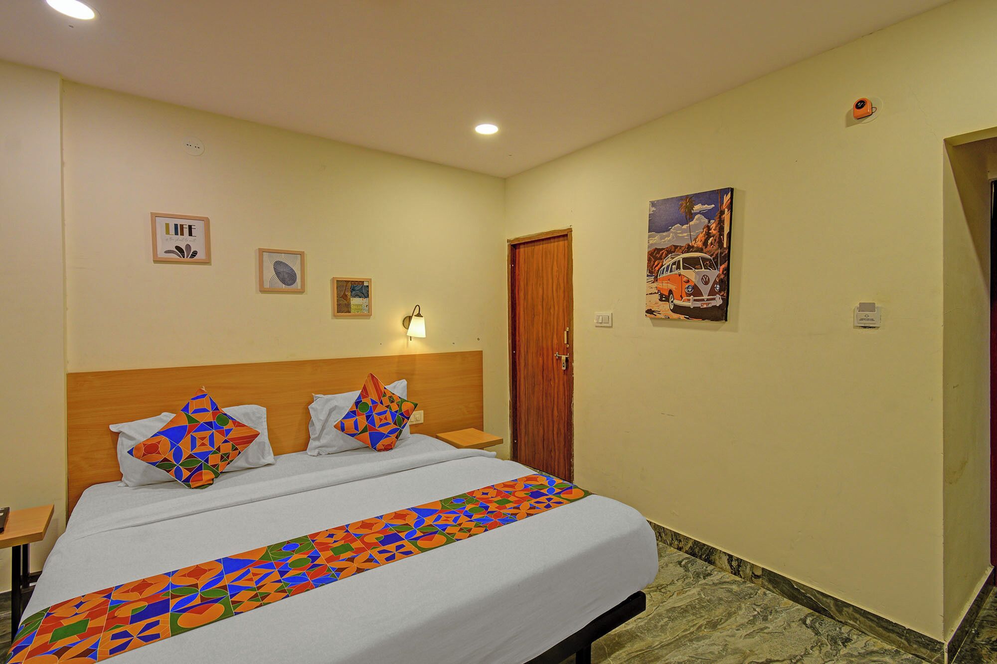 A hotel room with a bed, a wooden headboard, a wall-mounted lamp, and a door.
