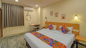 Una habitación de hotel con cama, escritorio, silla, ventilador de techo y aire acondicionado, además de una pared decorada con cuadros.