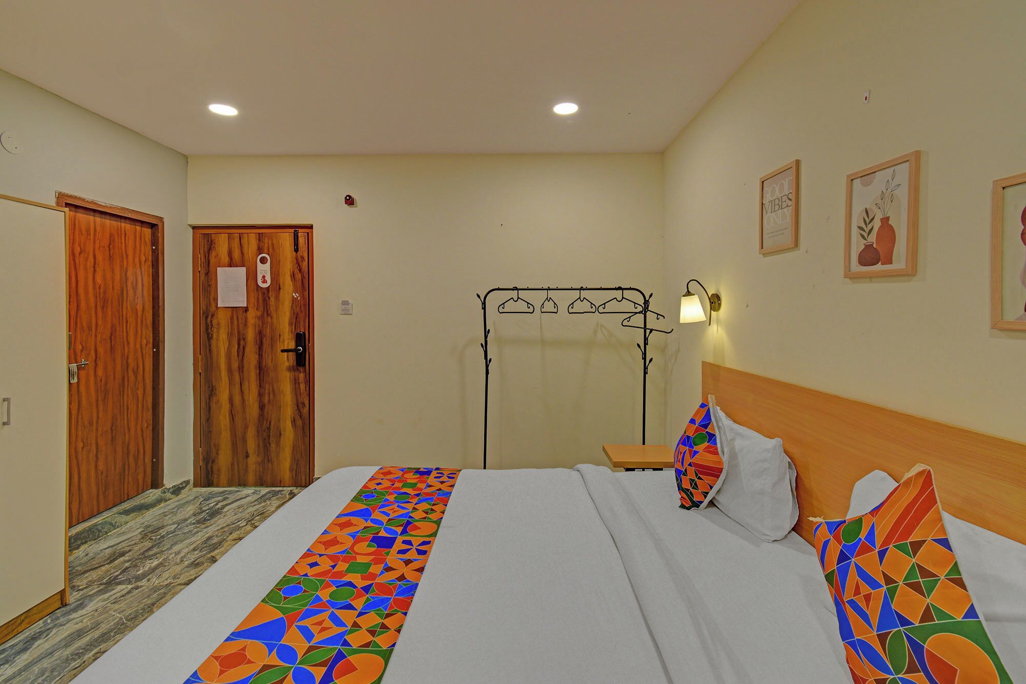A hotel room with a bed, a wooden door, a coat rack, and a wall with framed pictures.
