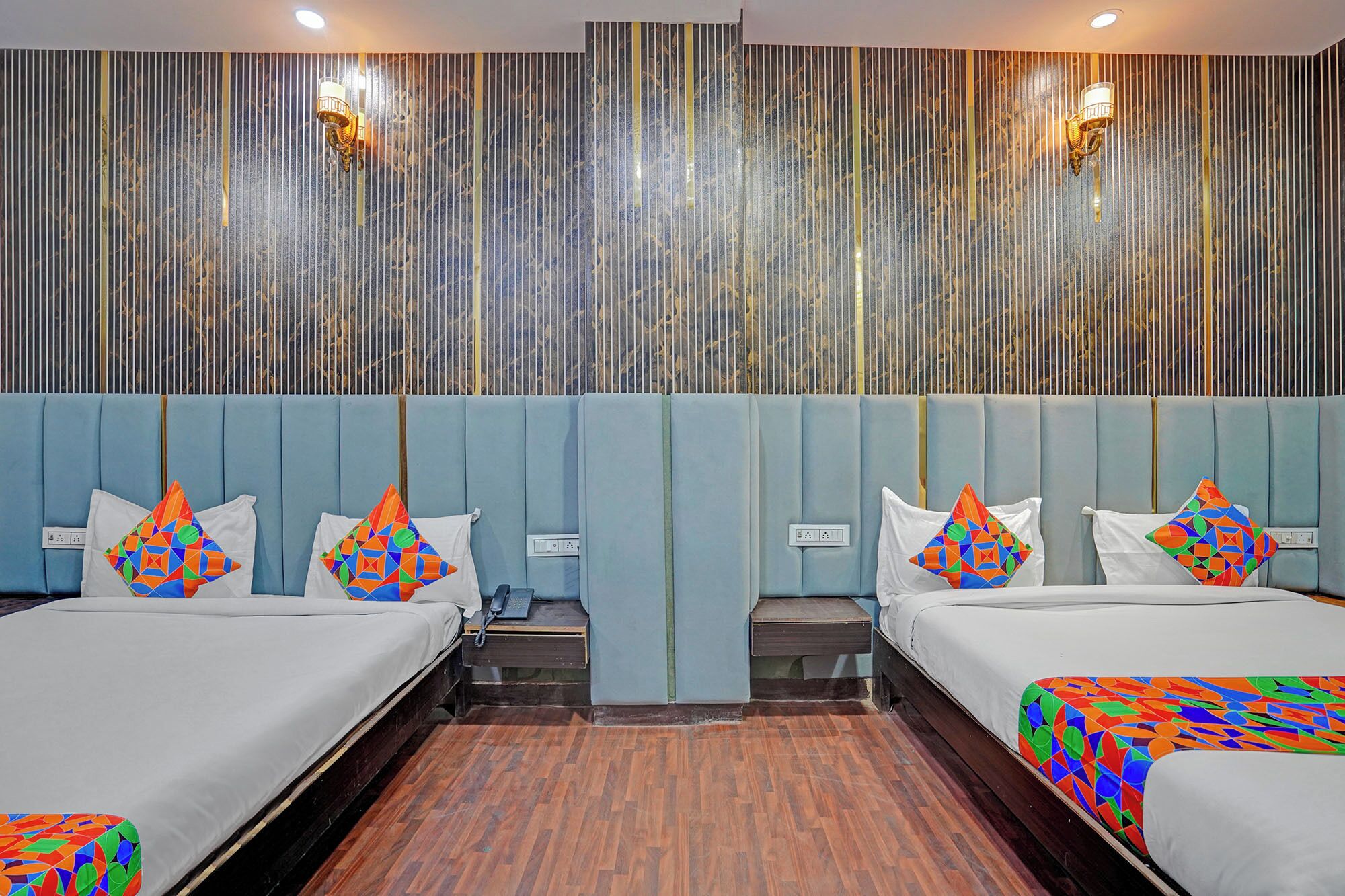 A hotel room with two beds, a wooden floor, a wall with vertical lines, and two wall-mounted lights.